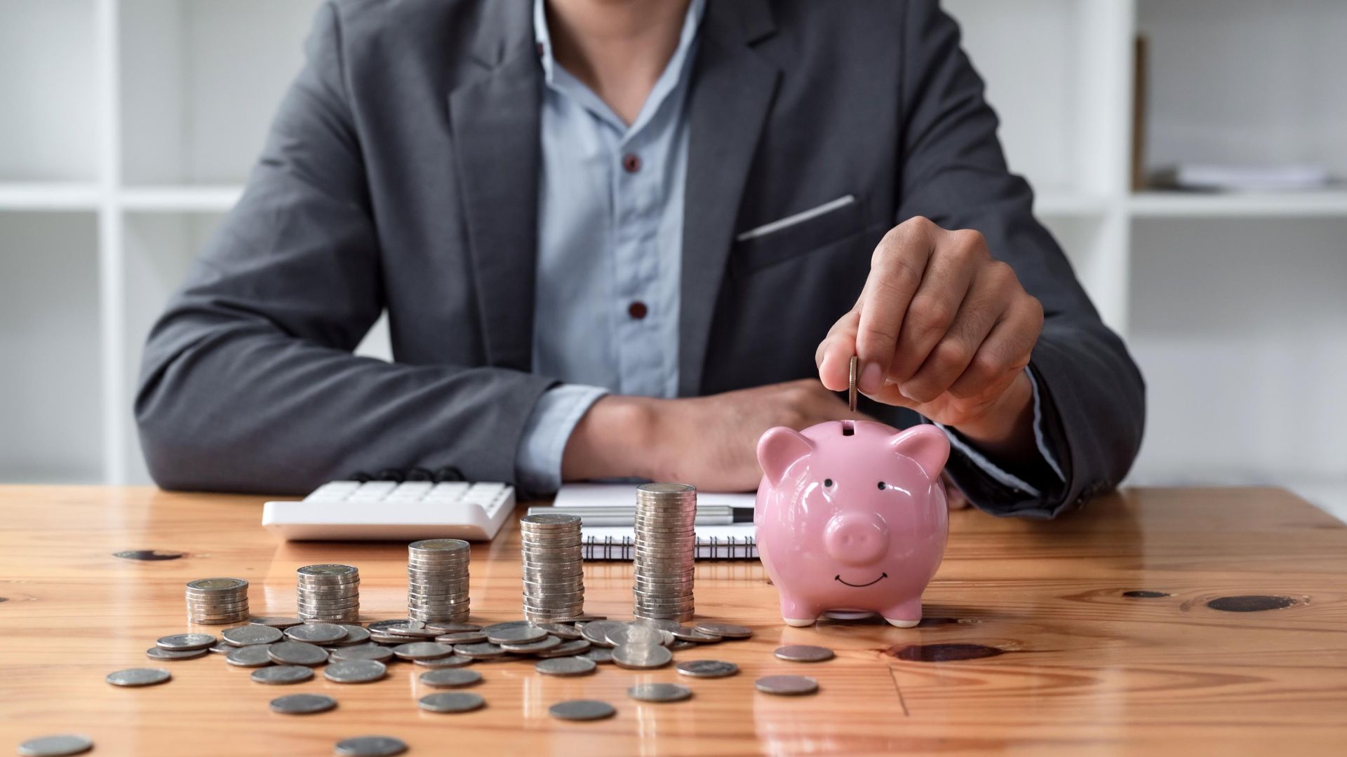 Person in suit putting coins into a pink piggy bank on a desk, with stacks of coins and a calculator.