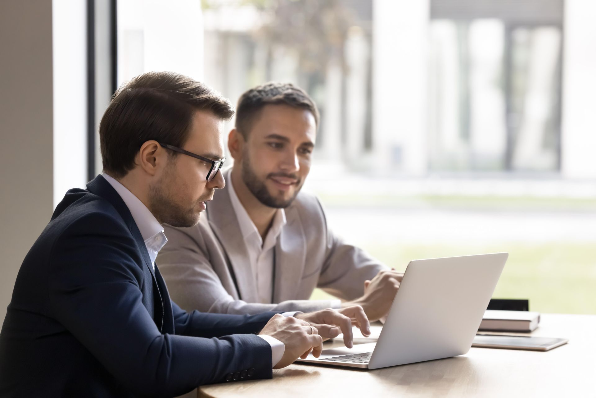 Two men in suits look at a laptop, one points and smiles, in a bright office.