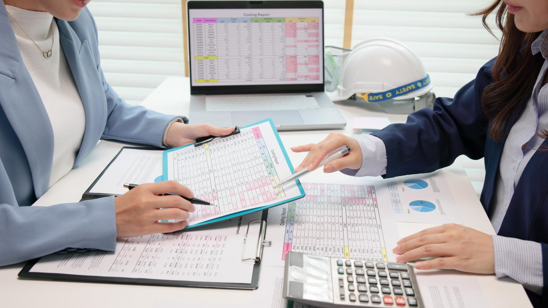 Two people reviewing documents at a desk, one pointing, a laptop, and a hard hat present.