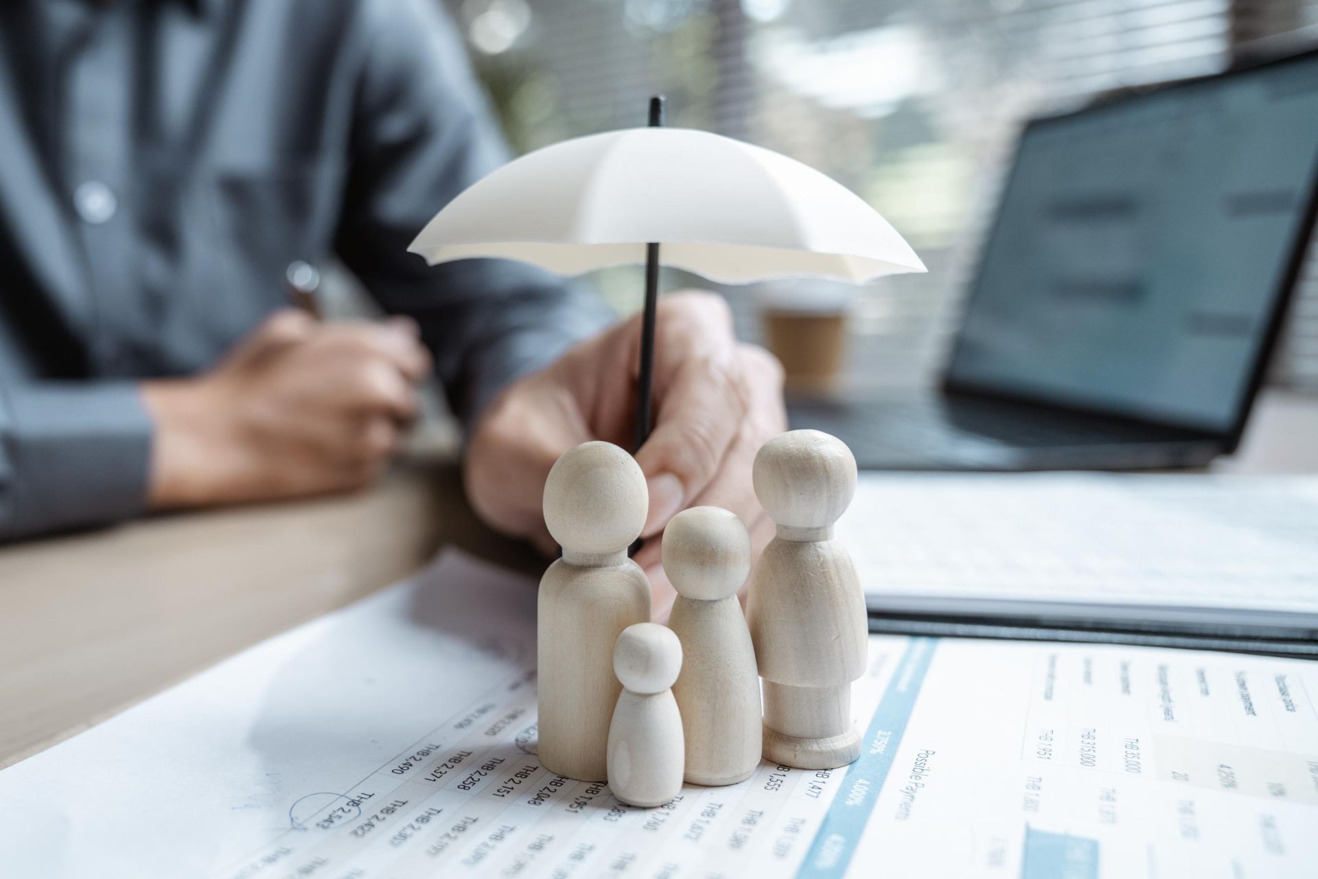 Wooden family figures under a small umbrella held by a person over paperwork and a laptop.