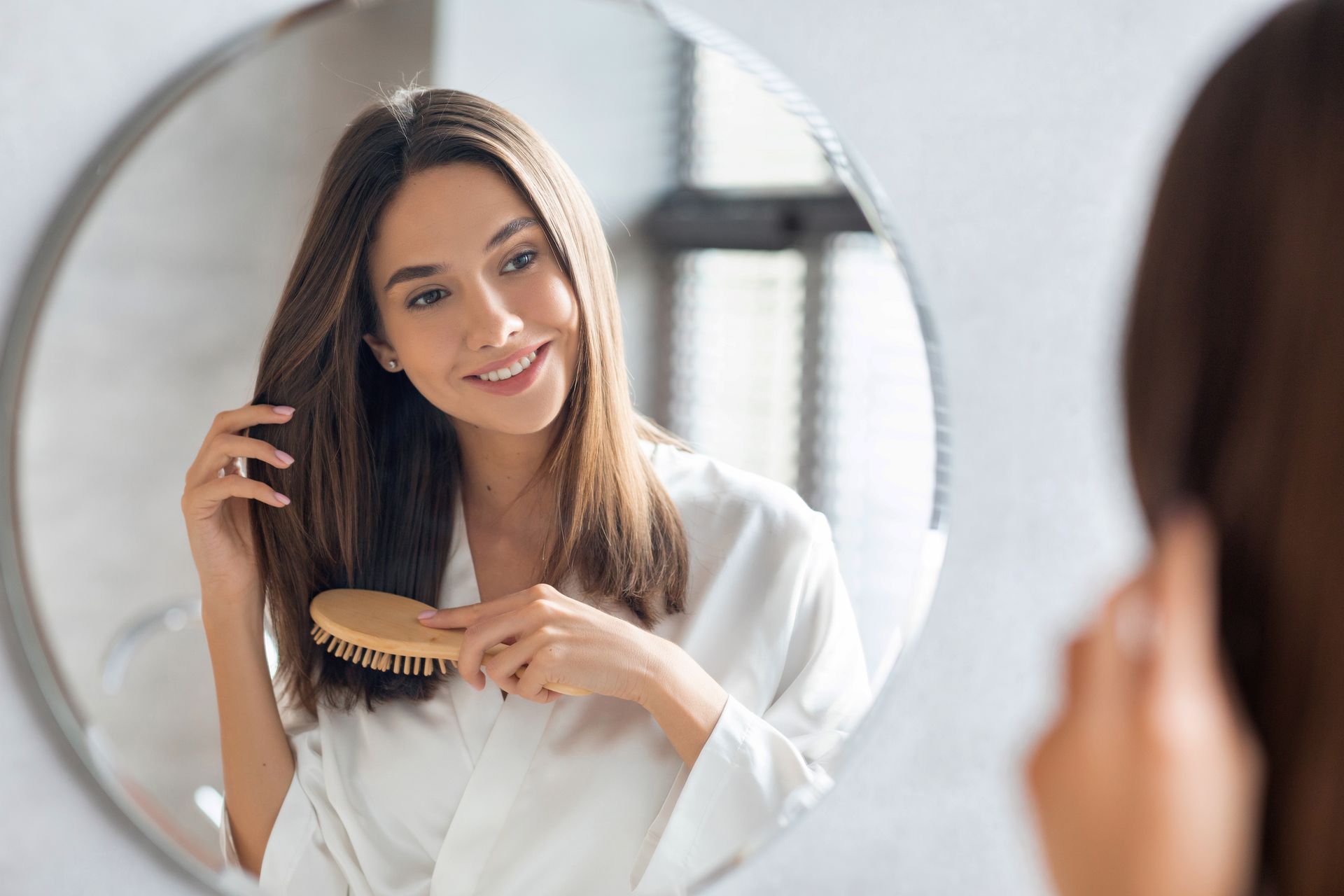 Woman in a white robe brushing her long brown hair in front of a mirror, smiling.