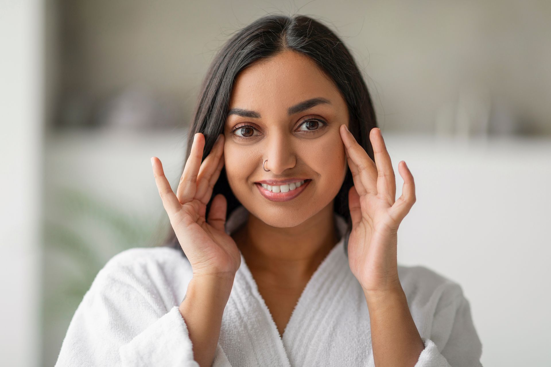 Woman in white robe smiles, gently touching her under-eye area.