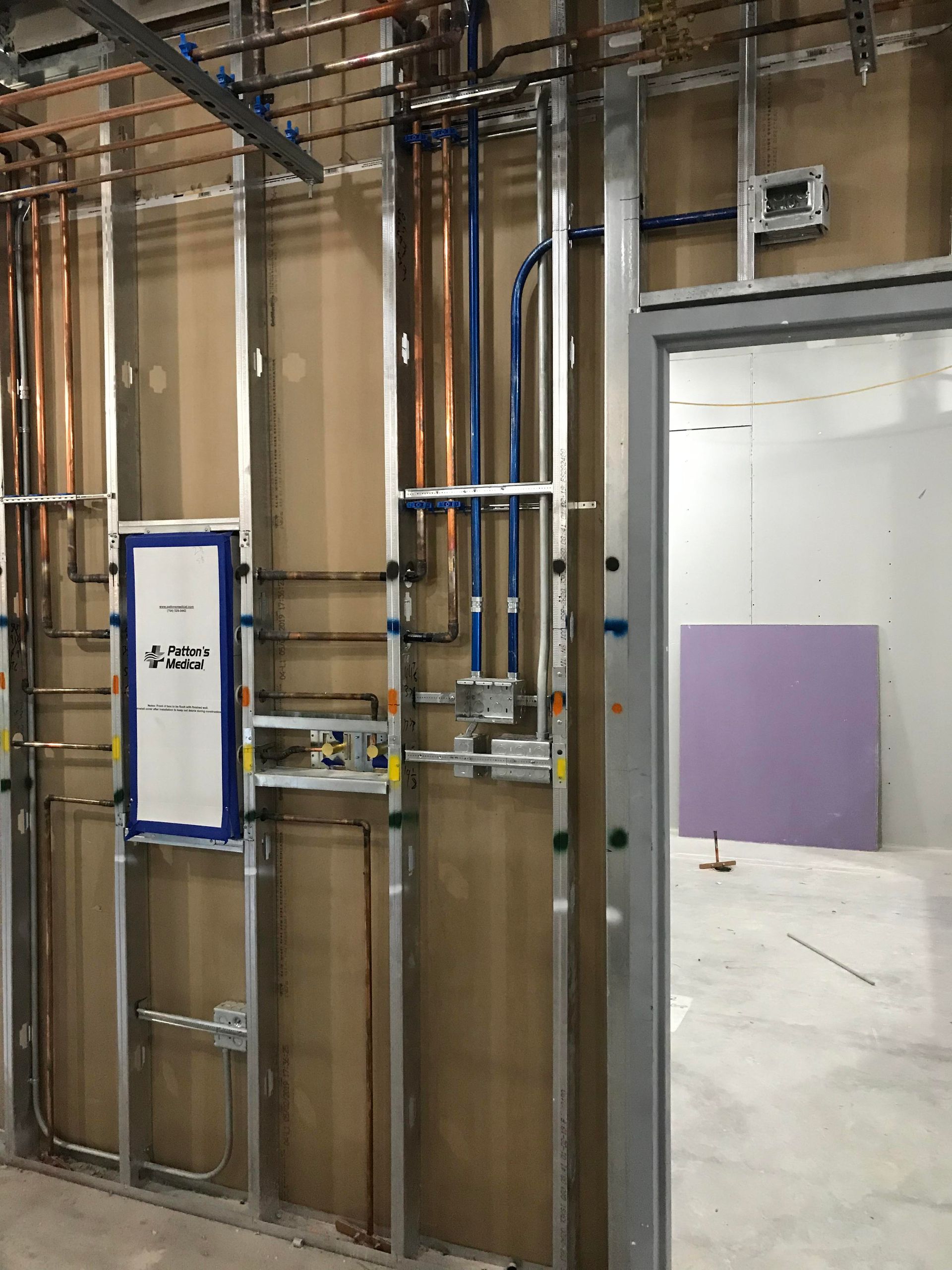 Interior view of unfinished construction, plumbing and electrical work visible within metal studs. Doorway leads to empty room.