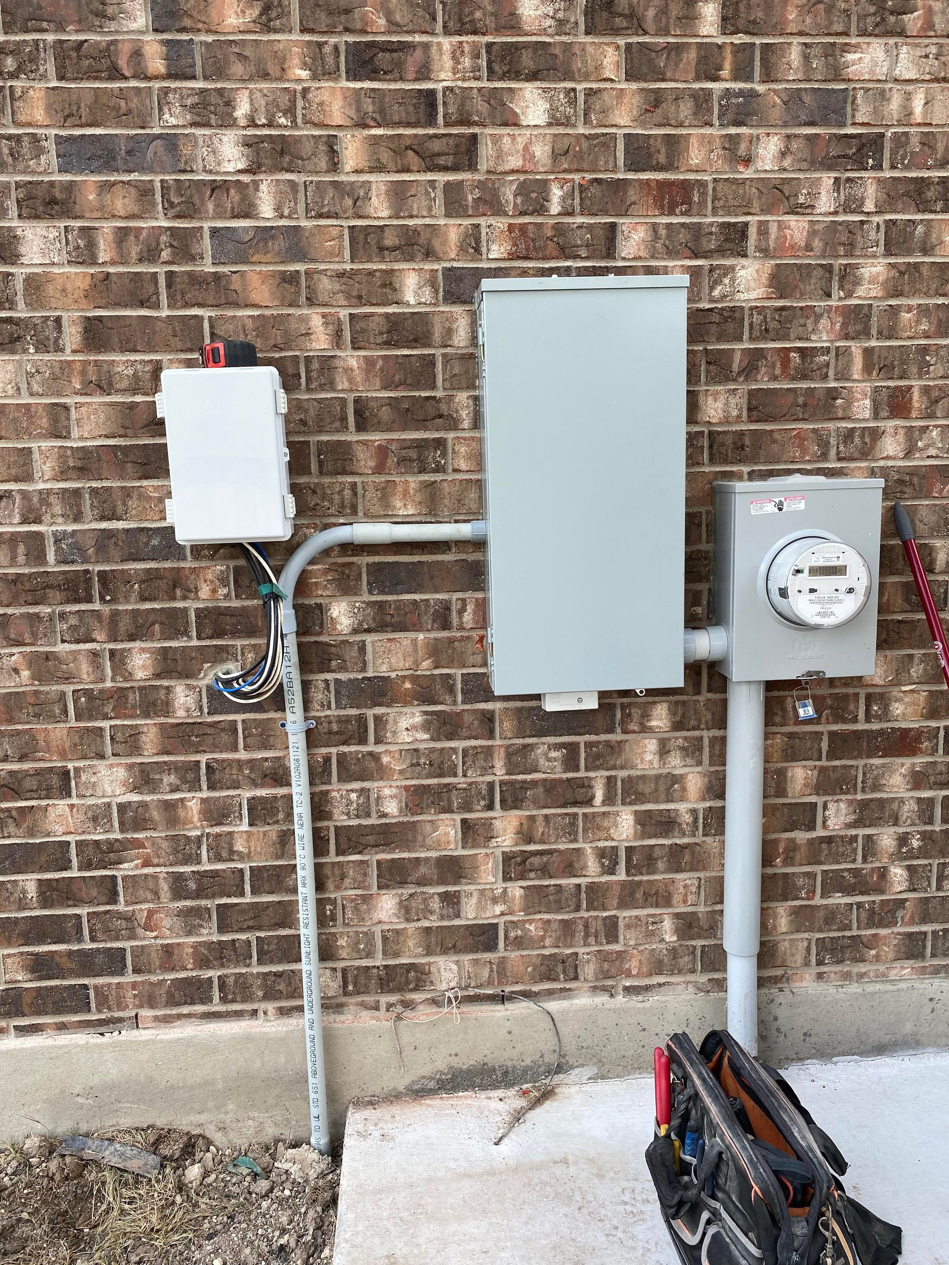 Exterior electrical panel and meter on a brick wall; a conduit connects the components.
