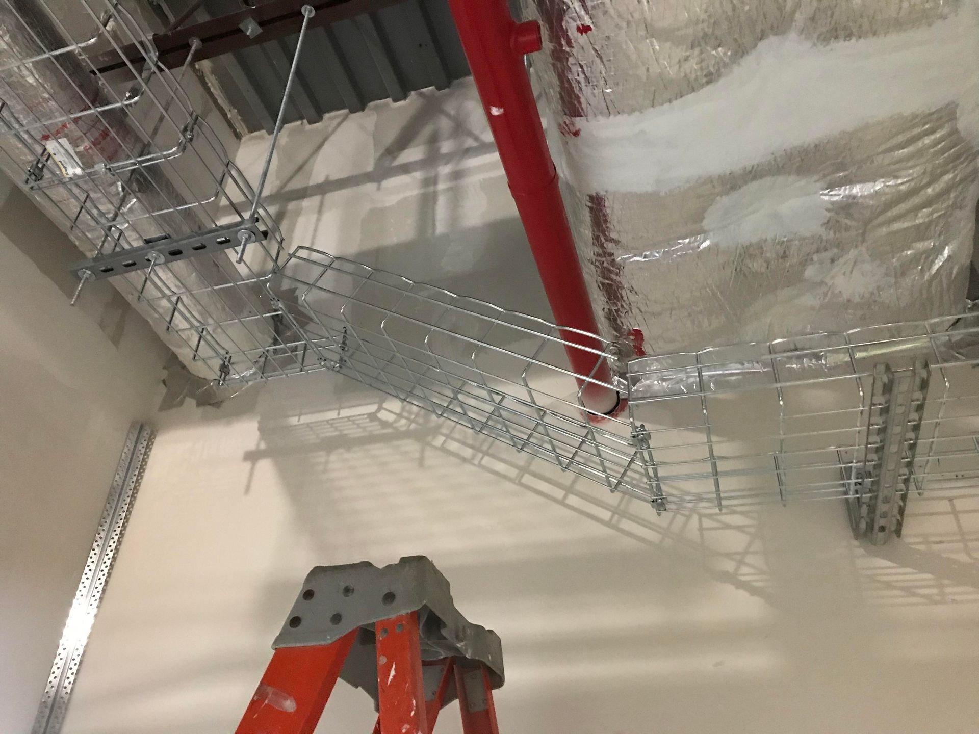 Ladder near a damaged metal cable tray and red pipe against a white wall.