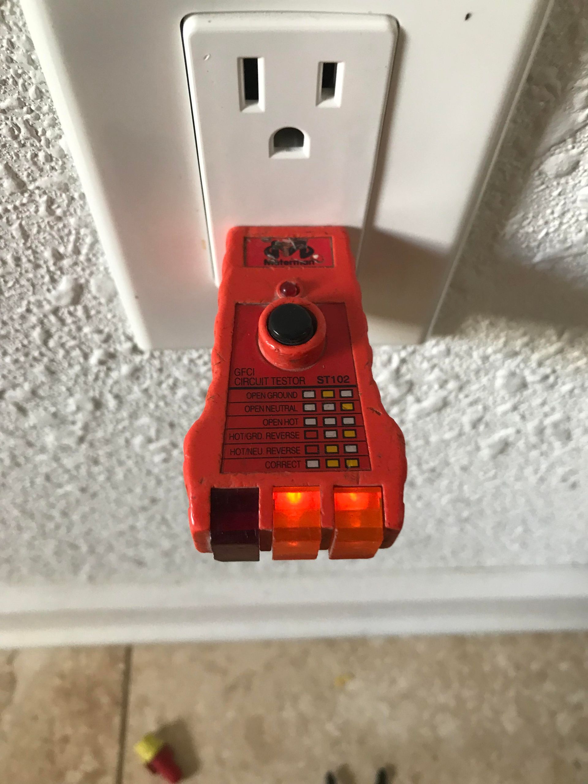 Red electrical outlet tester plugged into a white outlet, showing two orange and one red light, indicating a wiring fault.