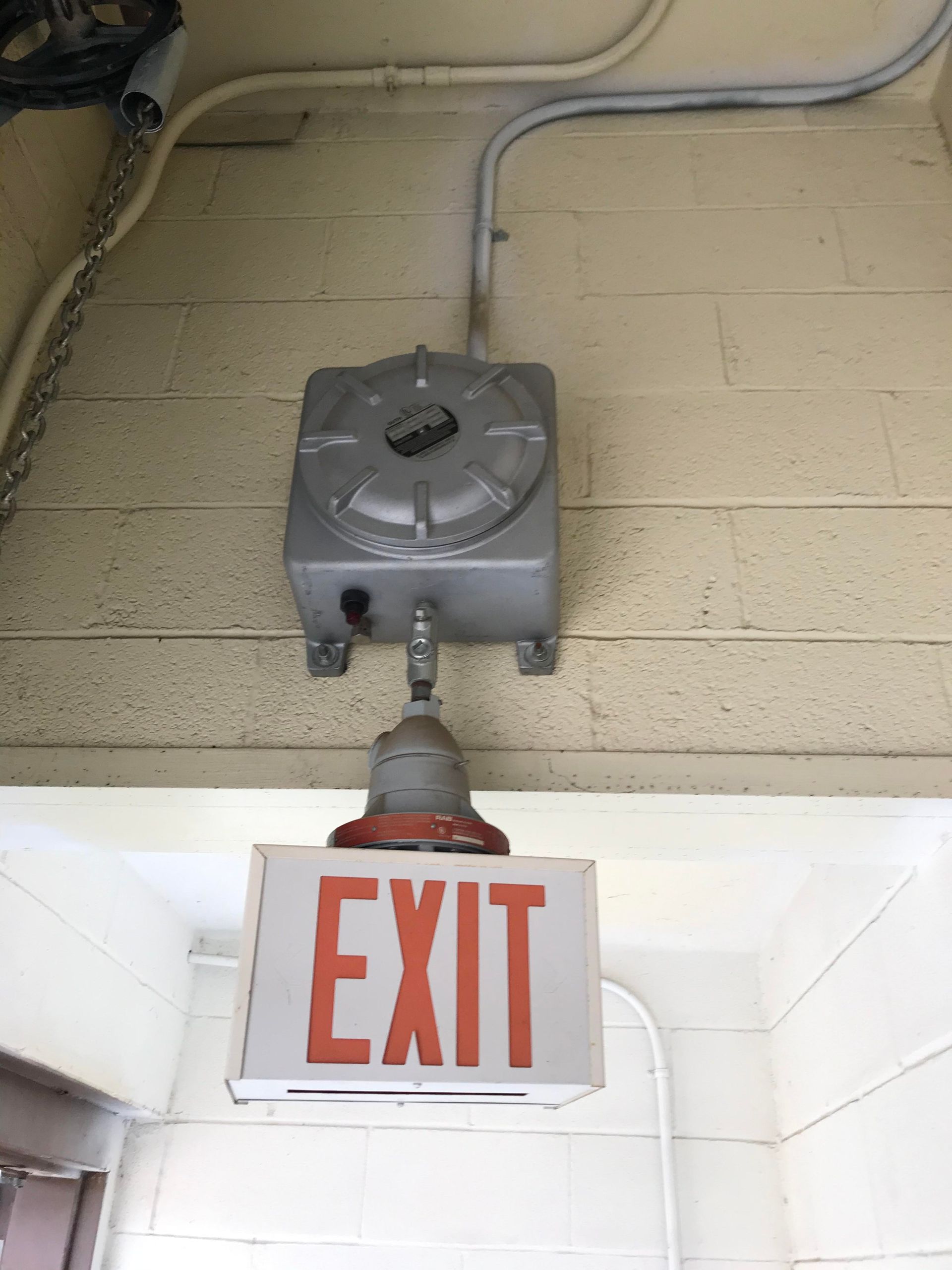 Exit sign below a gray metal mechanism on a light-colored brick wall.