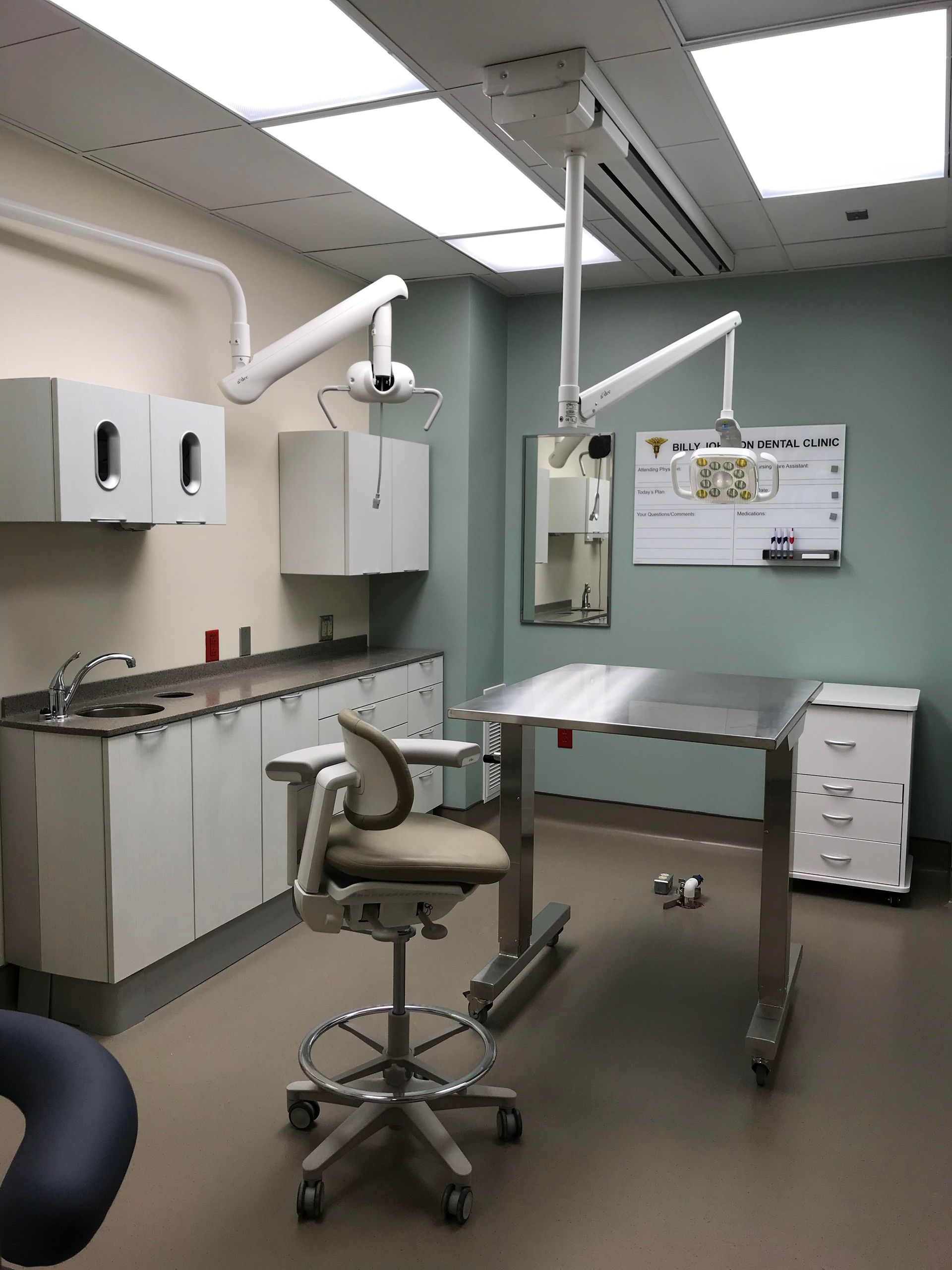 Exam room with stainless steel table, mounted lights, sink, cabinets, and a rolling chair.