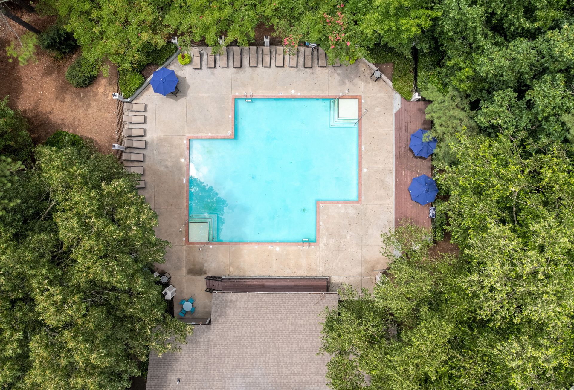 Aerial view of a rectangular, light blue swimming pool surrounded by a stone deck, lush green trees, and blue umbrellas.