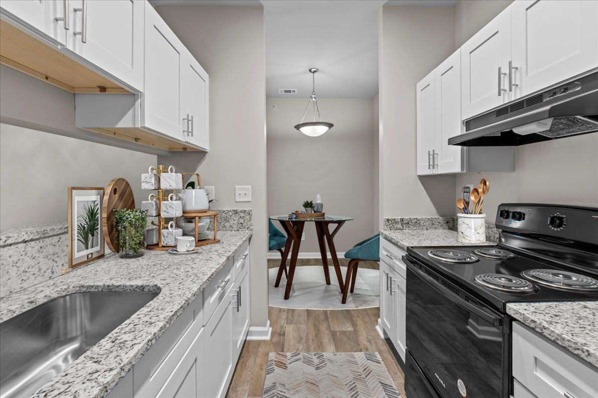 A galley kitchen with white cabinets, speckled countertops, a stainless steel sink, and a view into a dining area.