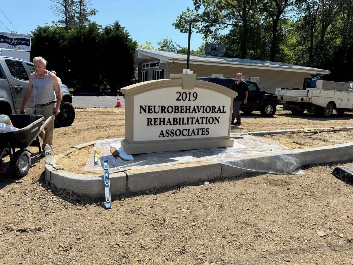 Stone sign for Neurobehavioral Rehabilitation Associates outside, with parked trucks and a person nearby.