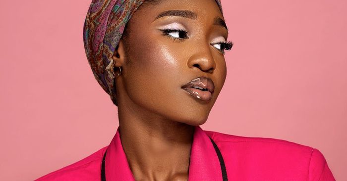 A woman wearing a head scarf and a pink jacket is standing in front of a pink background.