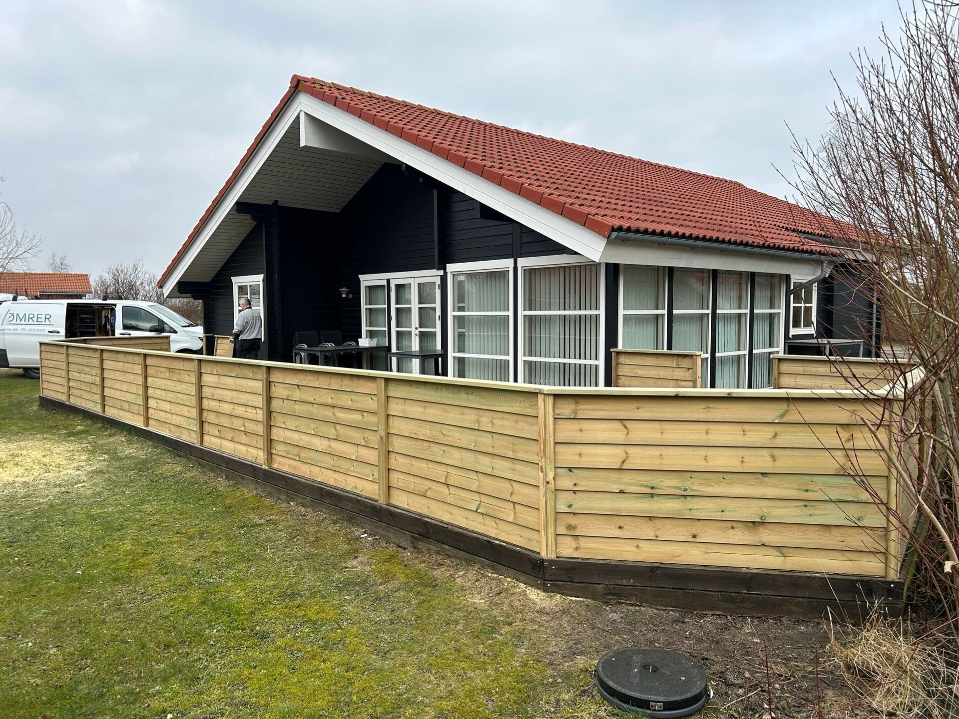 Et sort hus med rødt tag og et plankeværk foran.