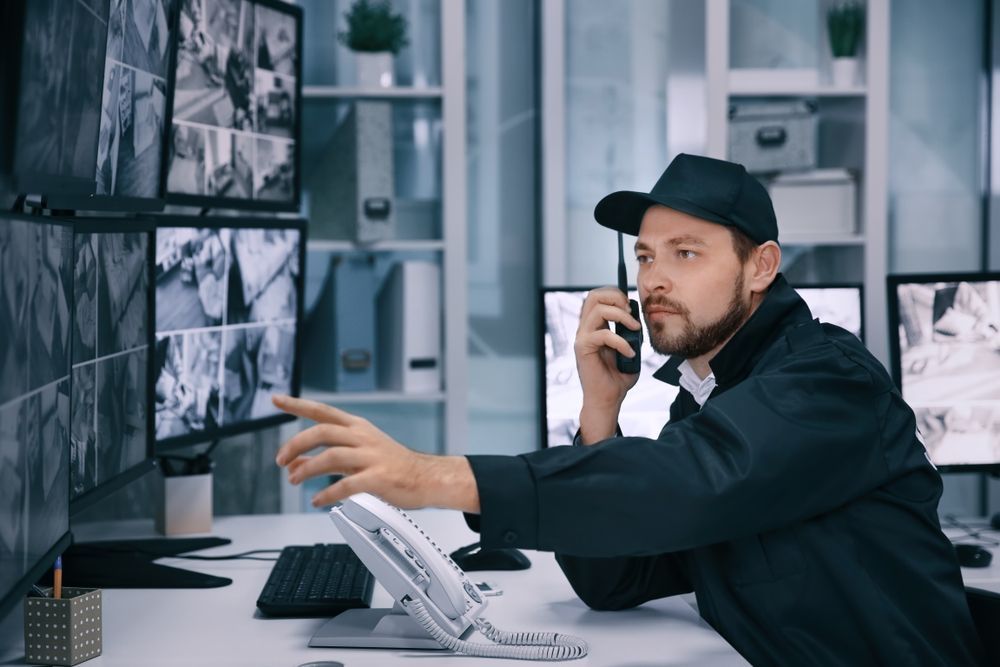 A security guard is talking on a cell phone while looking at a computer screen.