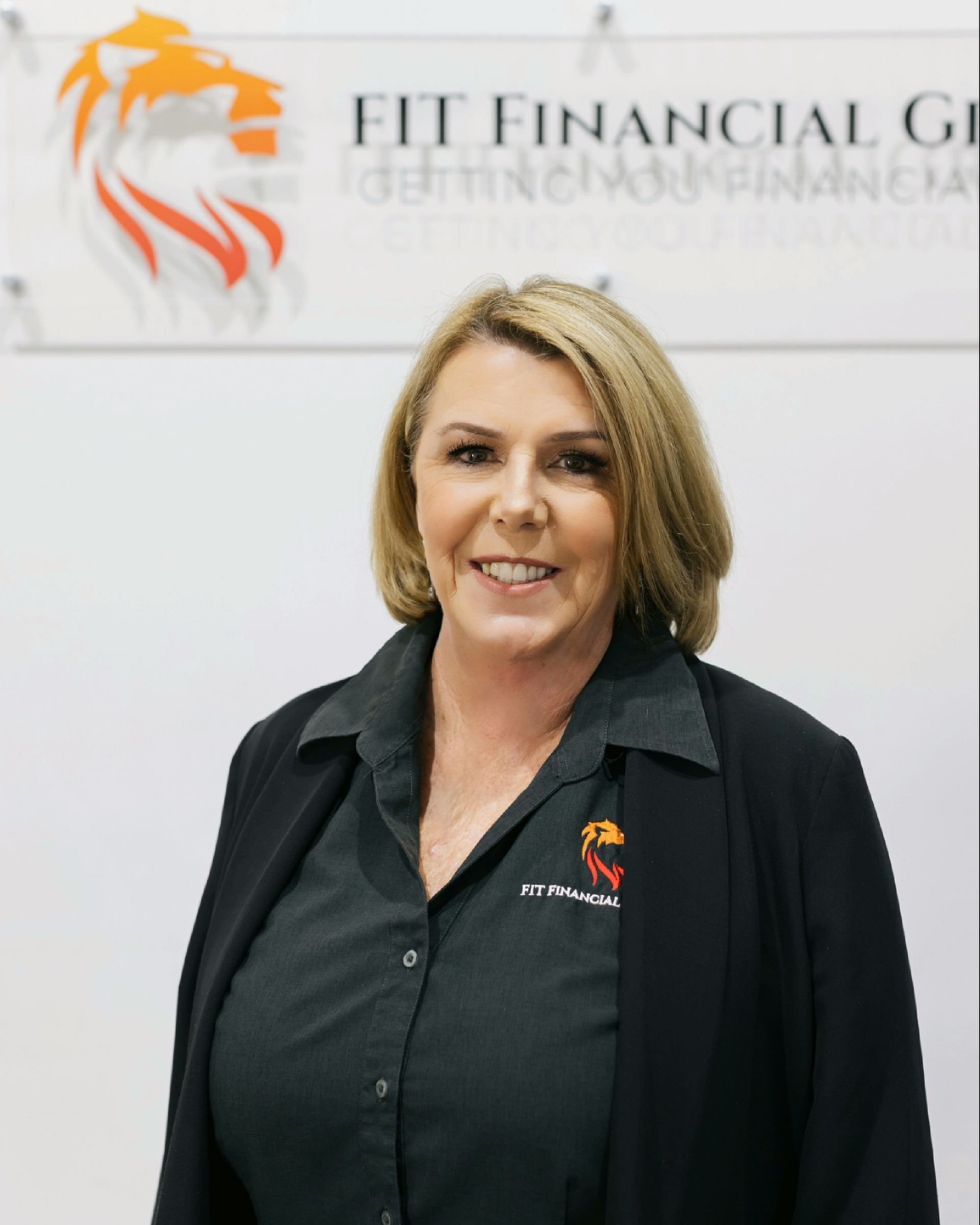 Woman in a black blazer and button-up shirt smiling in front of an office sign — Fit Financial Group in Leumeah, NSW