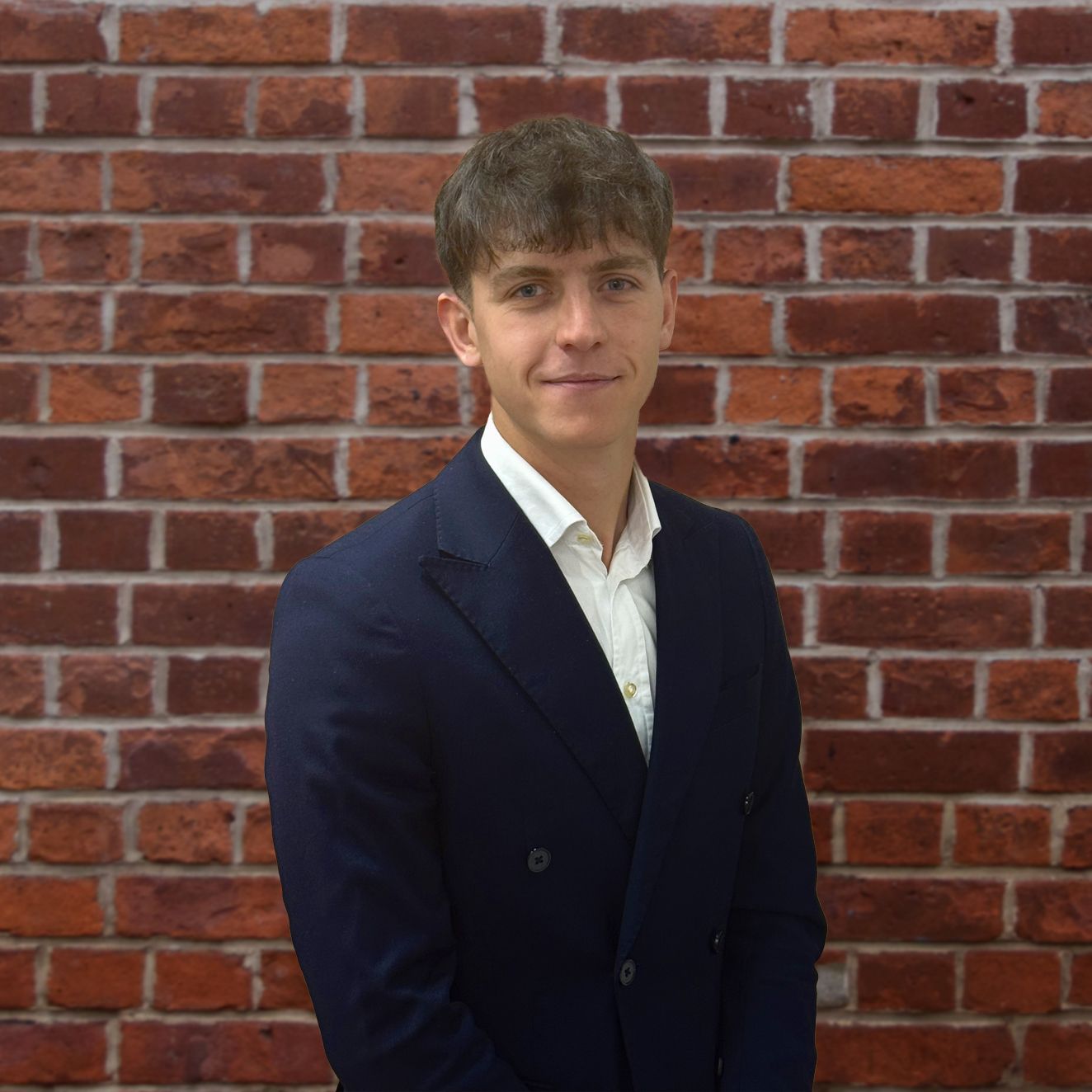 A young man in a suit and white shirt is standing in front of a brick wall.
