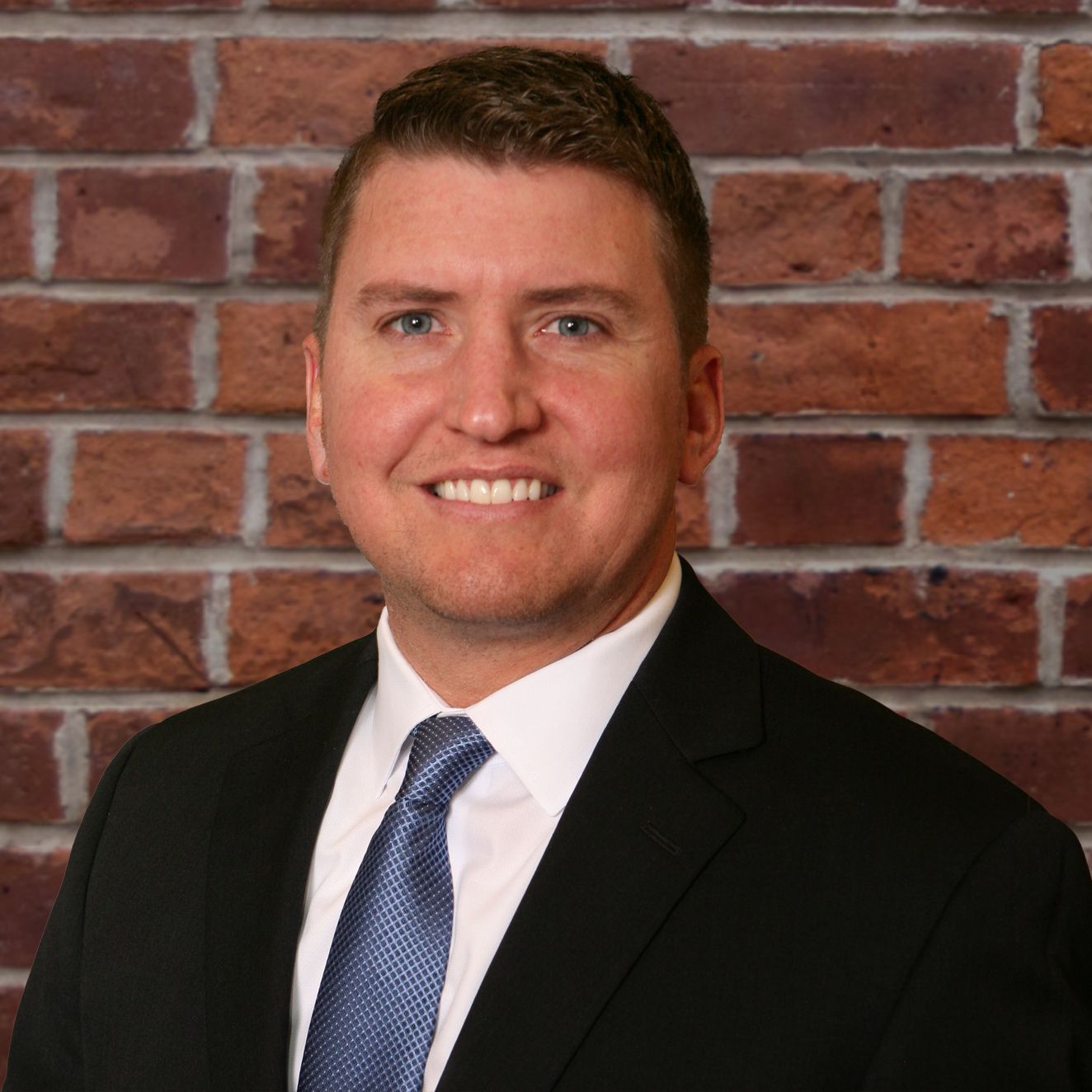 A man in a suit and tie is smiling in front of a brick wall.
