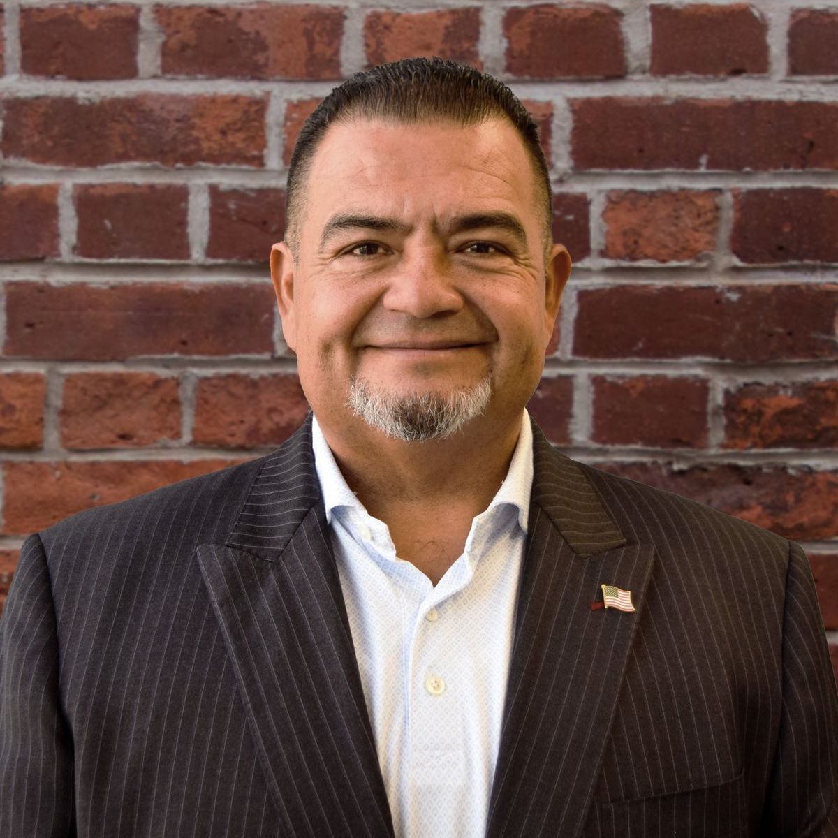 A man in a suit is smiling in front of a brick wall.
