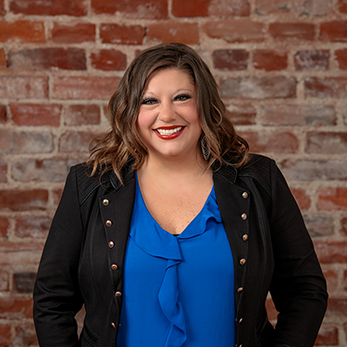 A woman in a blue shirt and black jacket is smiling in front of a brick wall.