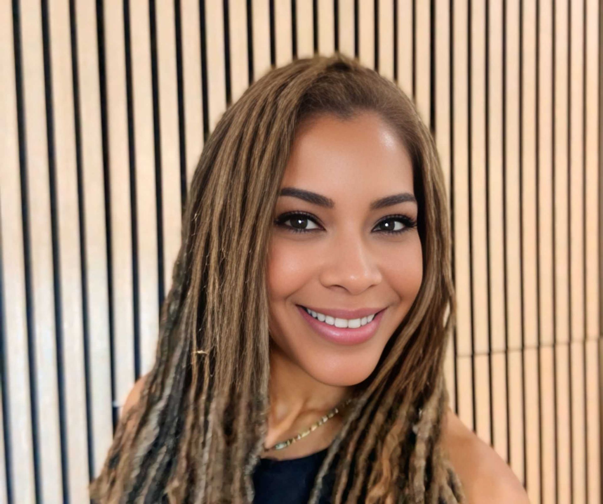 Woman with long, blonde dreadlocks smiles at the camera against a striped background.