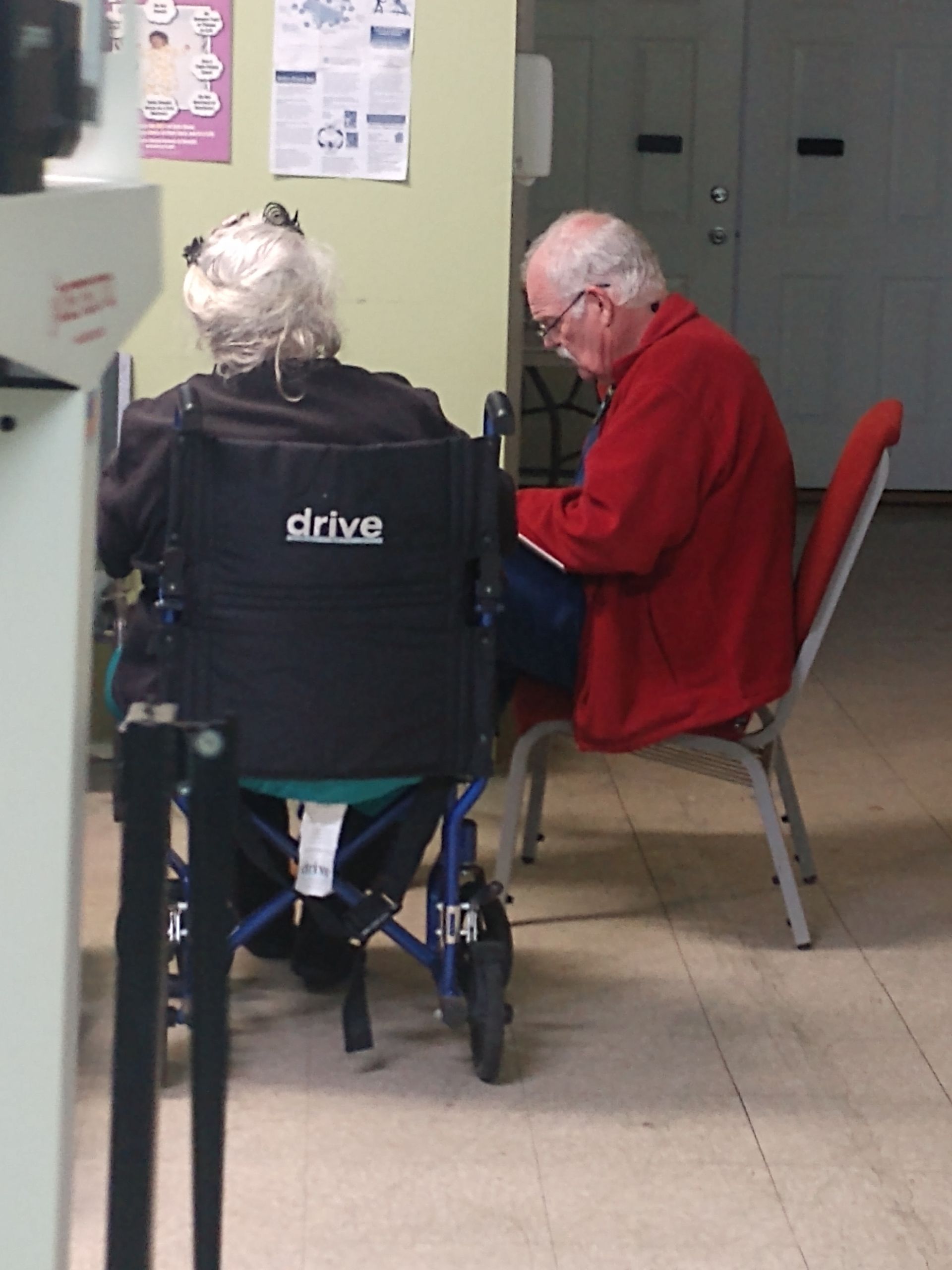 A man in a red jacket sits next to a woman in a black drive wheelchair