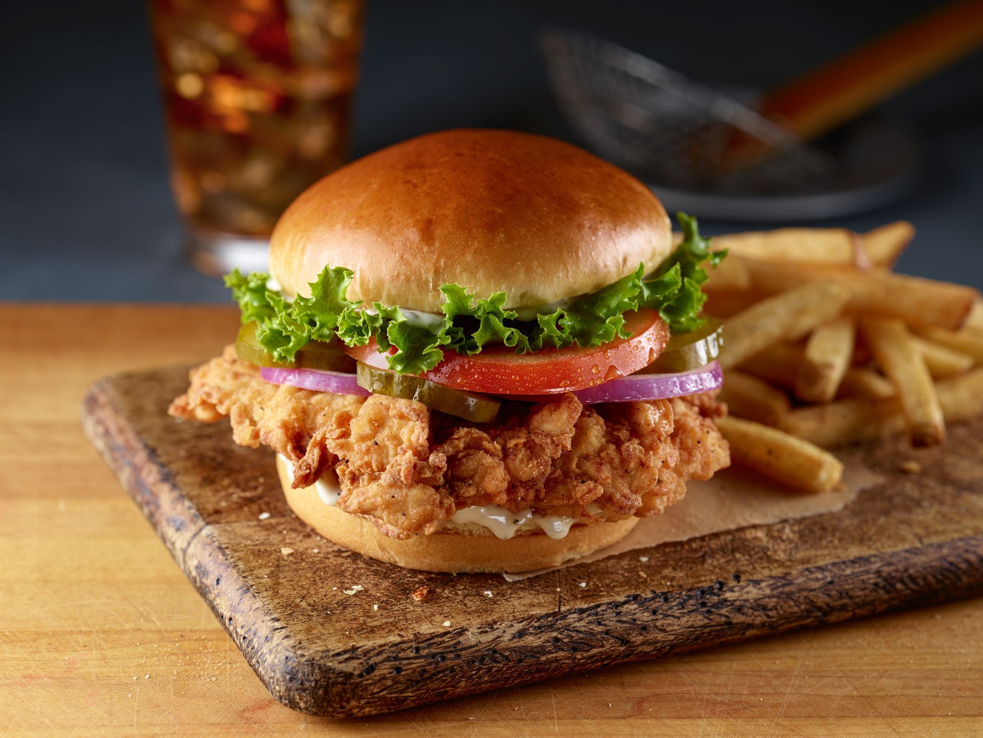 A crispy fried chicken sandwich with lettuce, tomato, and red onion on a wooden board with a side of golden french fries.