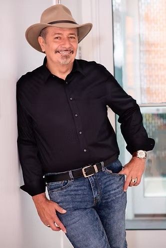Man in black shirt, jeans, and cowboy hat leaning against a wall, smiling.