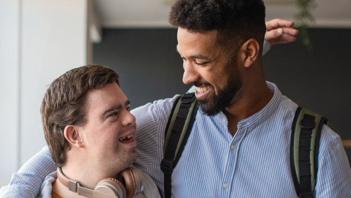 A man with a backpack is hugging another man with a disability.