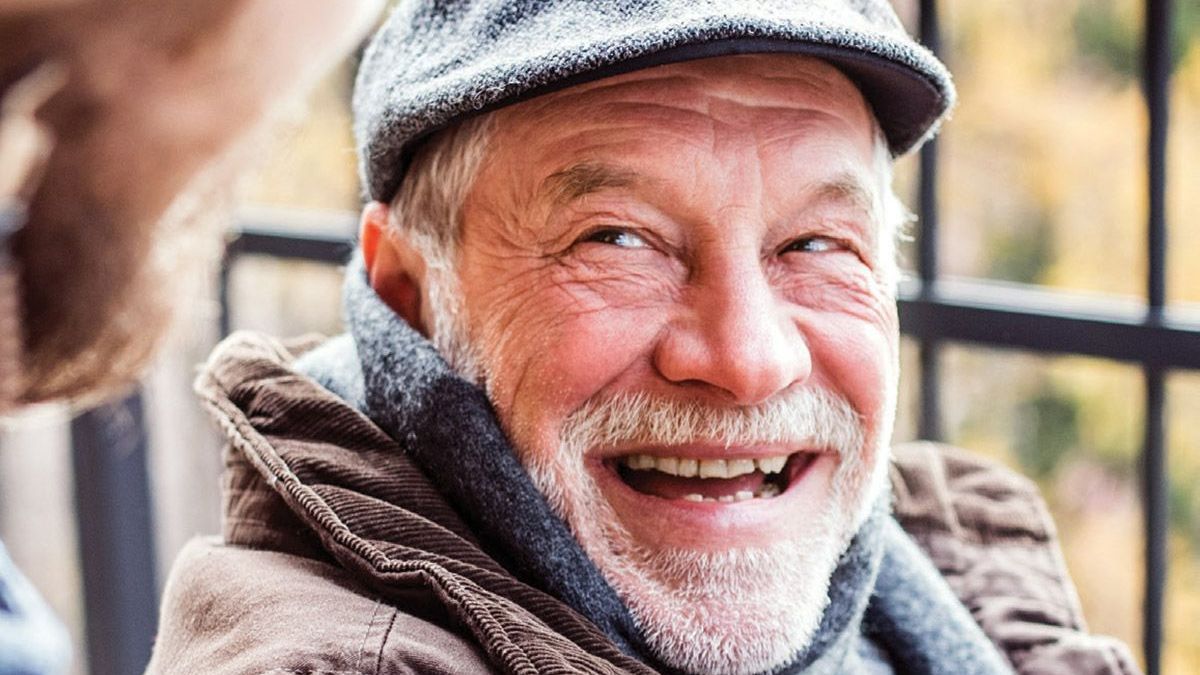 An elderly man wearing a hat and scarf is smiling.