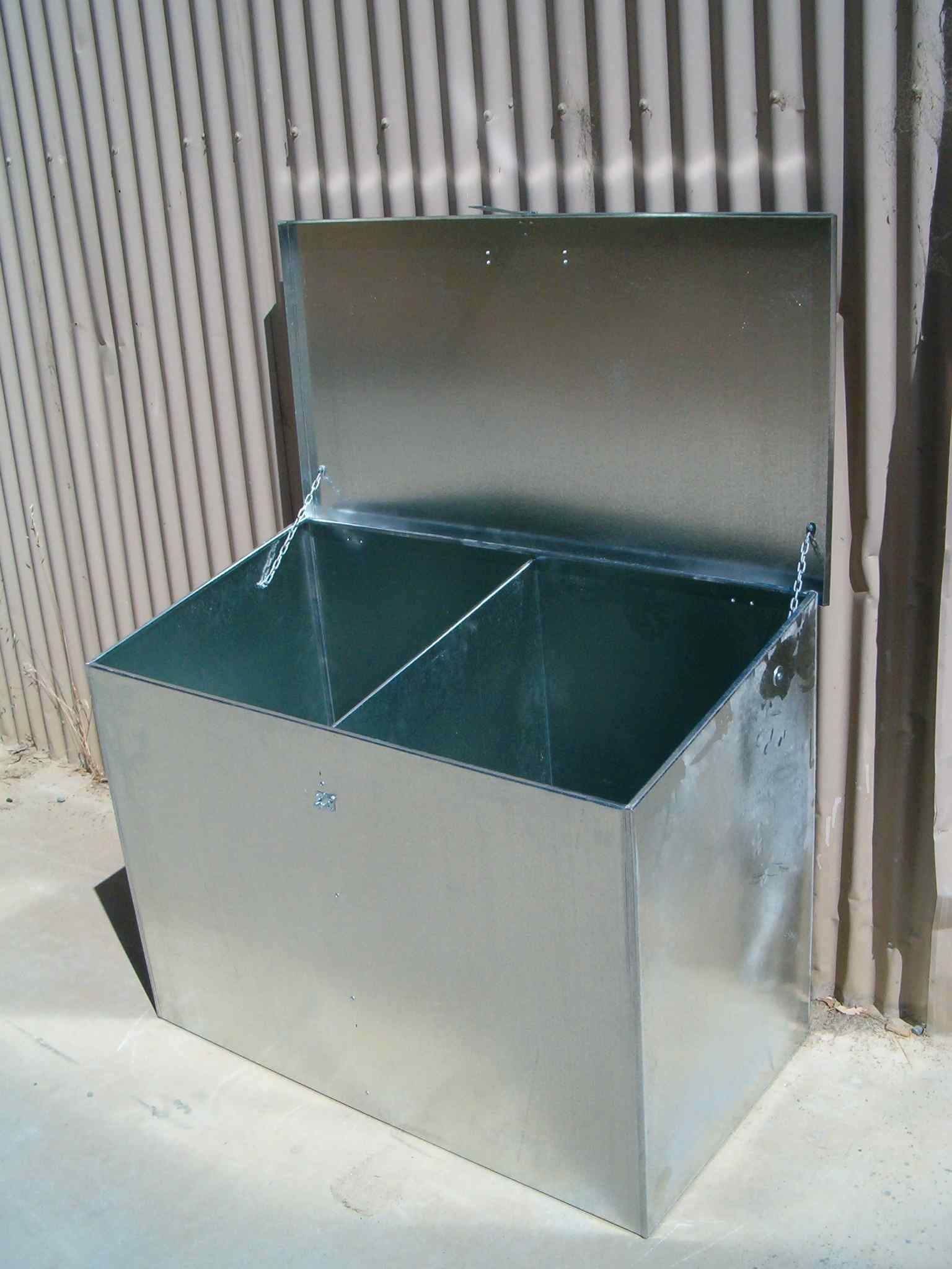 A metal box with the lid open is sitting in front of a fence.