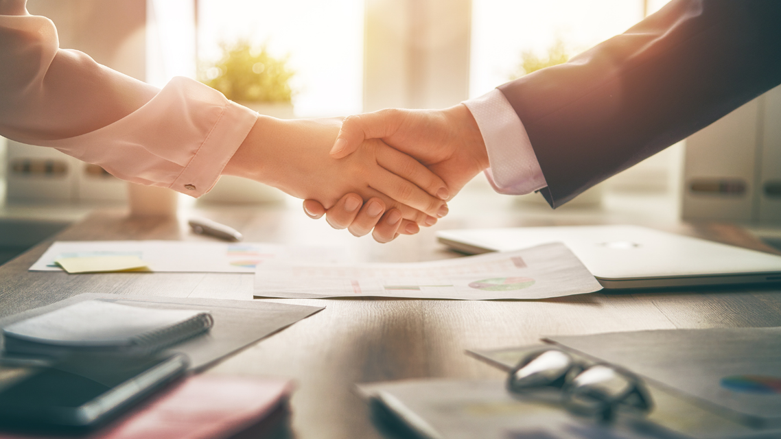 Two people shake hands over business documents on a desk in a bright, sunlit office.