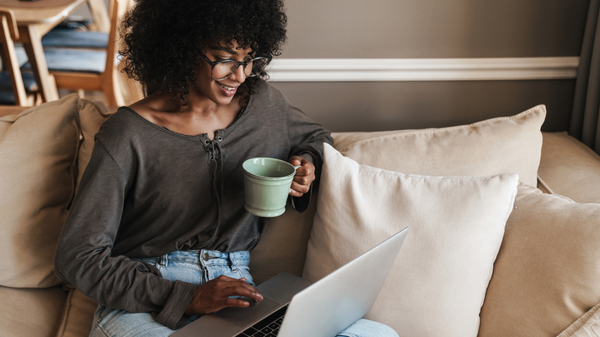 A person with curly hair smiles while sitting on a couch, holding a mug and using a laptop.