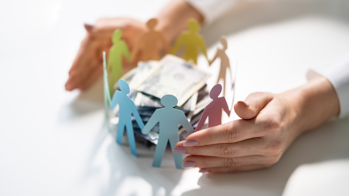 Hands surround a circle of colorful paper cutout people standing around a stack of US dollar bills on a white surface.