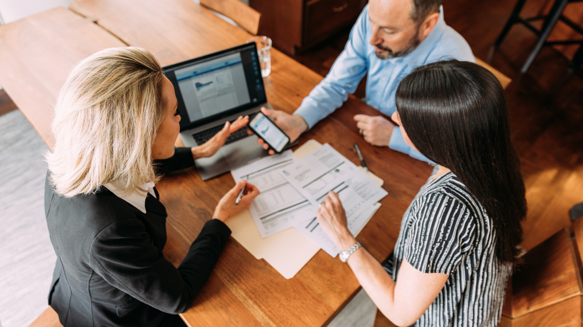 Three professionals sitting at a table with a laptop and documents, collaborating on a project in a modern office space.