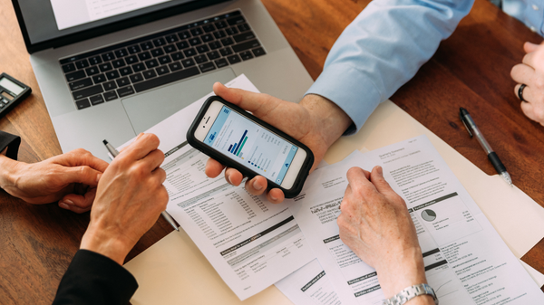 Three people review financial documents and a smartphone screen on a wooden desk with a laptop and a pen nearby.