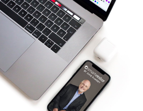 Laptop, wireless earbuds, and a smartphone displaying the SmartVestor by Dave Ramsey app on a white surface.