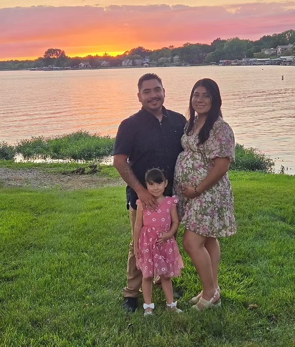 A pregnant woman , a man and a little girl are standing in front of a body of water.