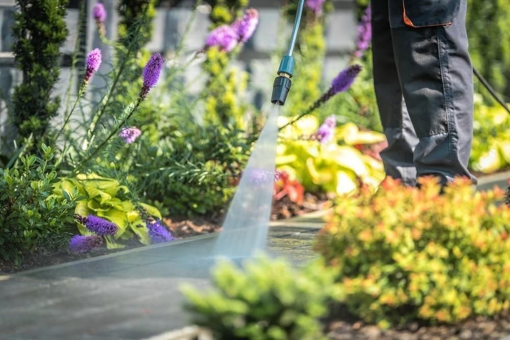 A person is using a high pressure washer to clean a sidewalk in a garden.