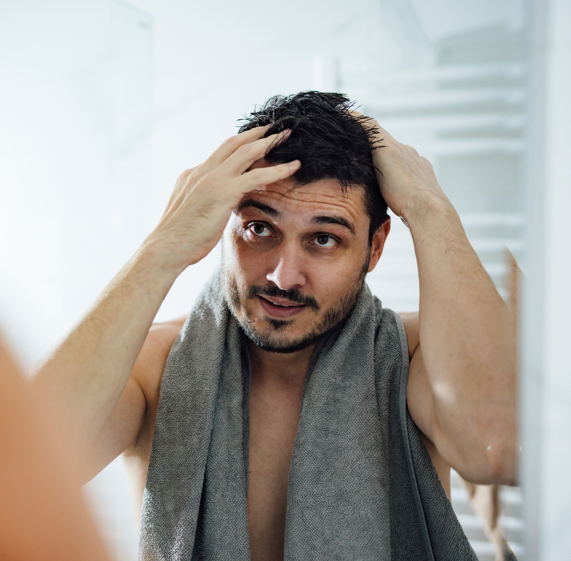 A man with a towel around his neck is looking at his hair in the mirror.
