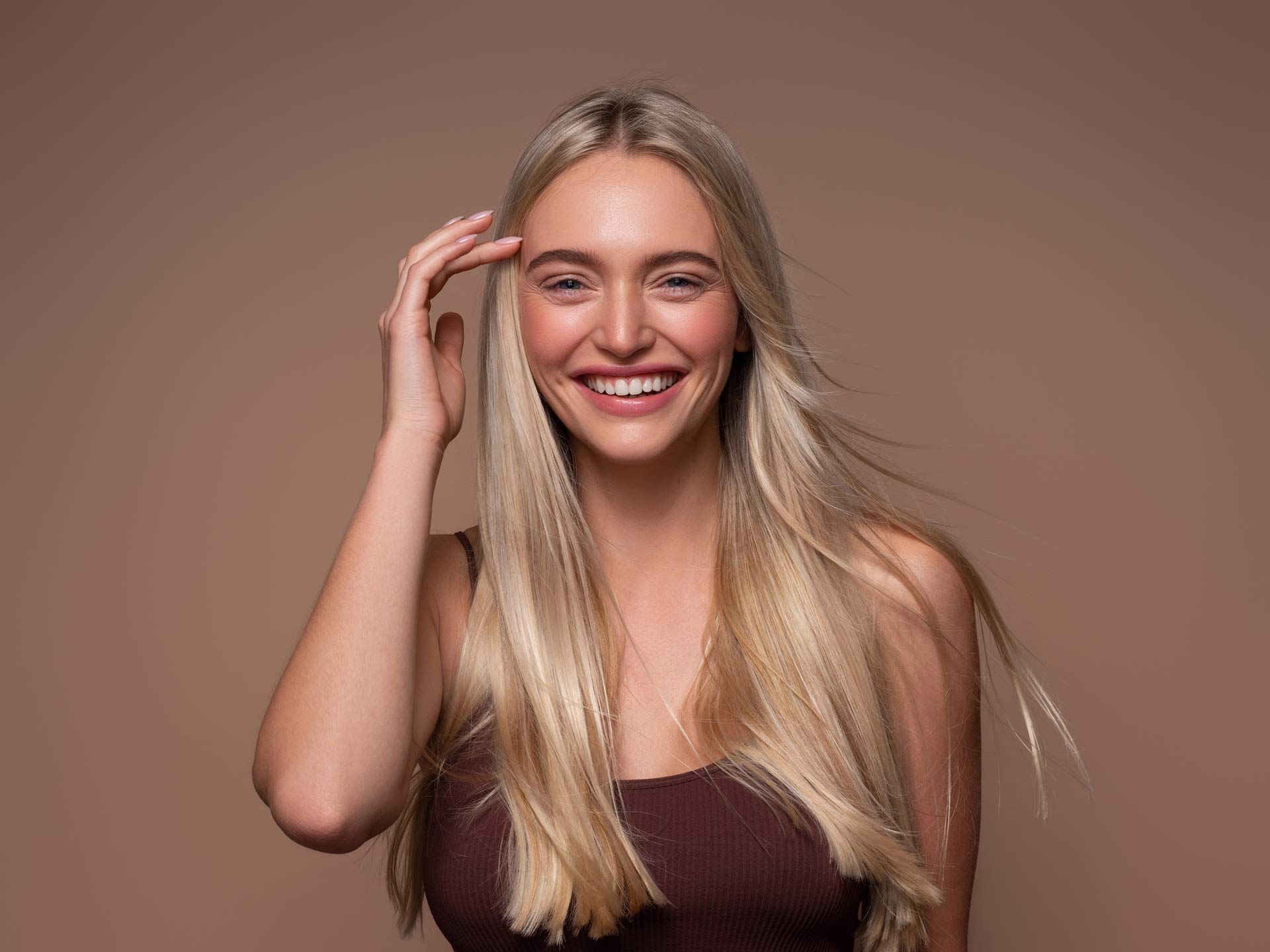 A woman with long blonde hair is smiling and touching her hair.