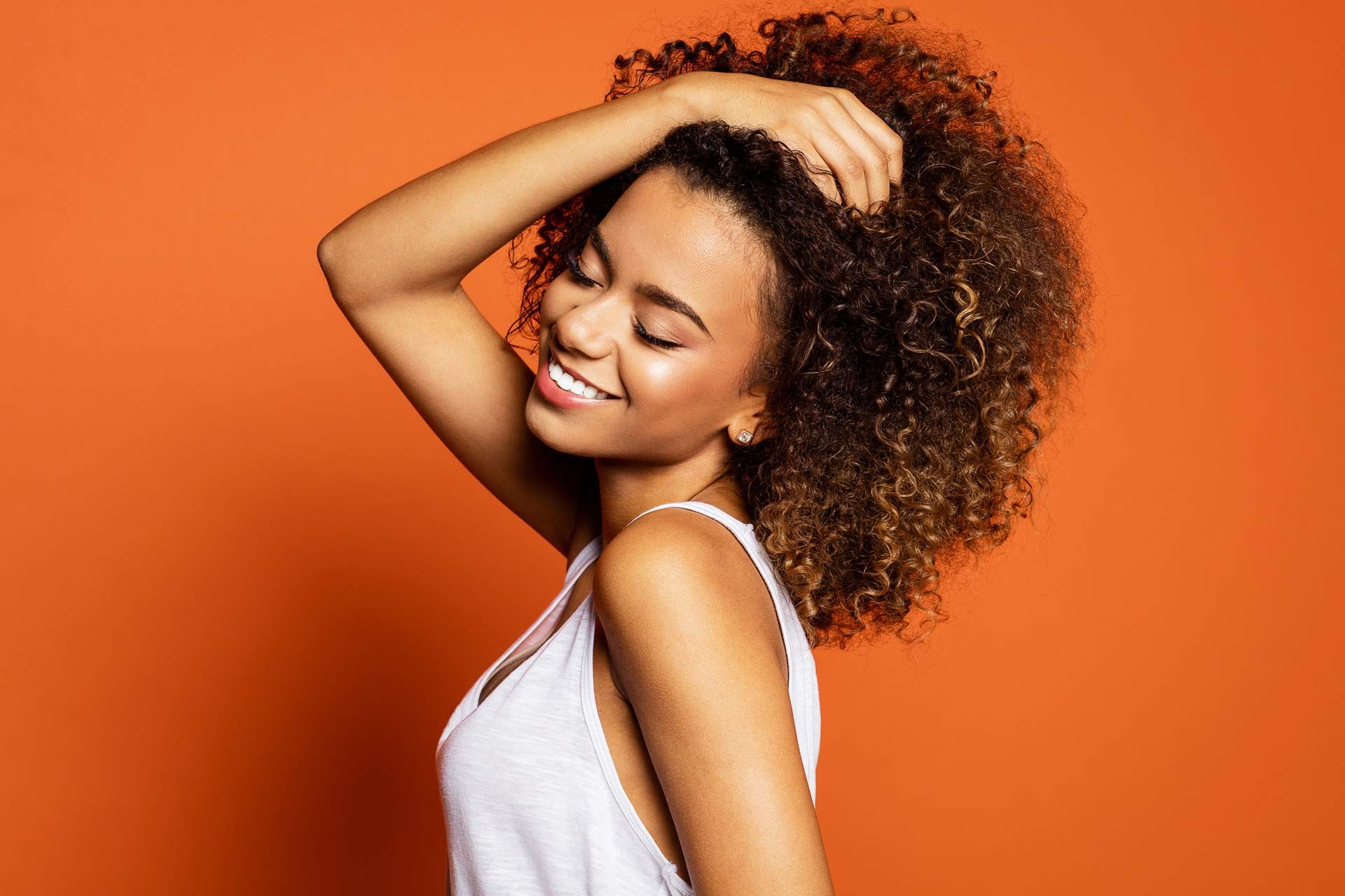 A woman with curly hair is smiling and touching her hair.