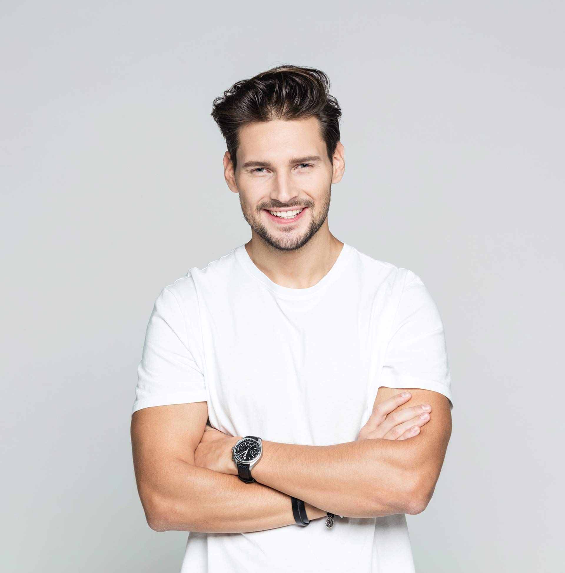 A man wearing a white t-shirt and a watch is standing with his arms crossed and smiling.