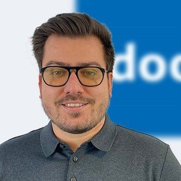 Ein Mann mit Brille lächelt vor einem blauen Schild mit der Aufschrift „Doc“