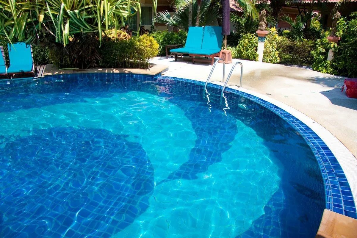 Swimming pool with blue tiles and water, surrounded by concrete and greenery.