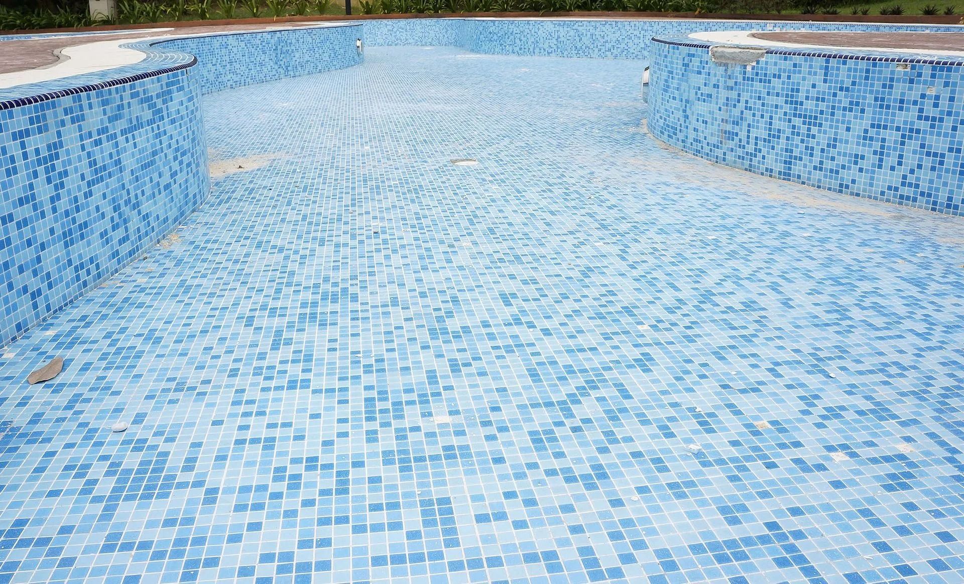 Empty blue-tiled swimming pool. The pool is dry and has some bare concrete patches.