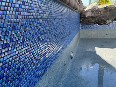 Blue mosaic tile interior of a pool with gray concrete and a rock feature.