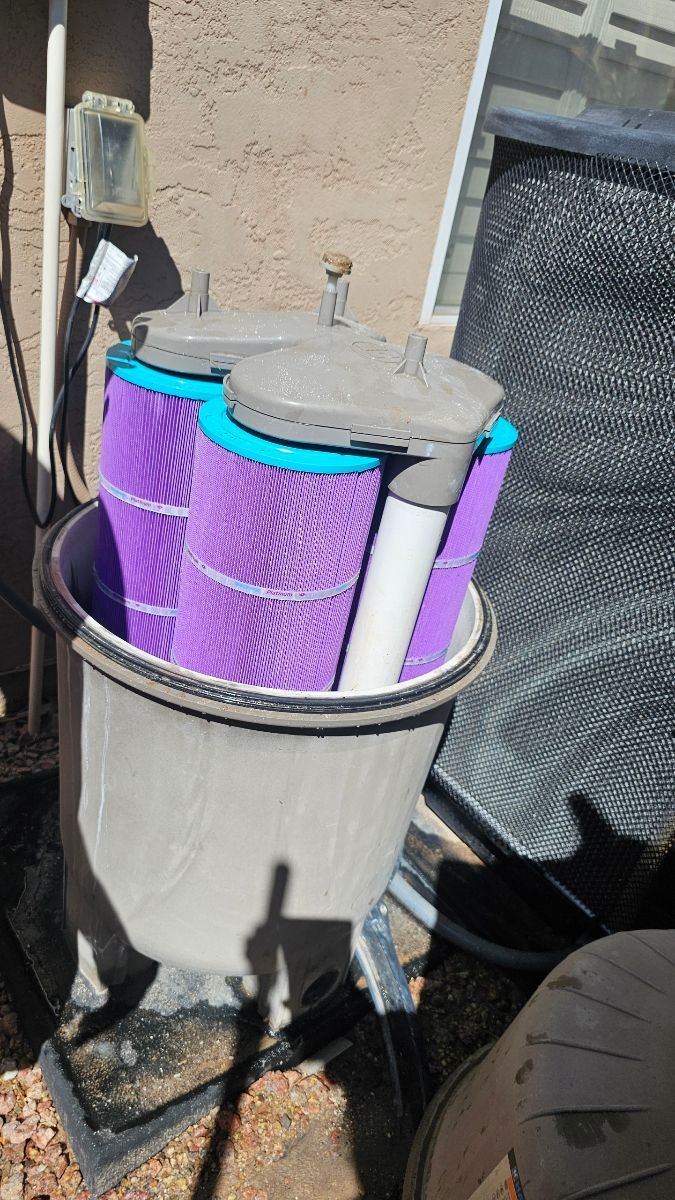 Outdoor view of a pool filter with three purple filter cartridges inside a gray tub.