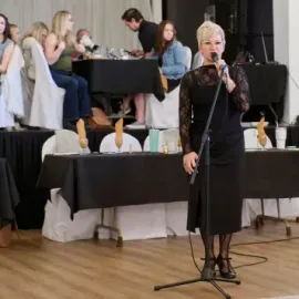 Woman speaking into a microphone at a banquet table setup in a hall