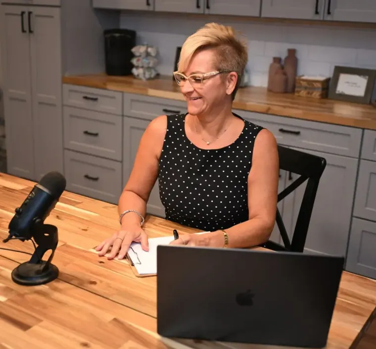Woman speaking at a wooden table with a microphone and laptop in a modern kitchen office area