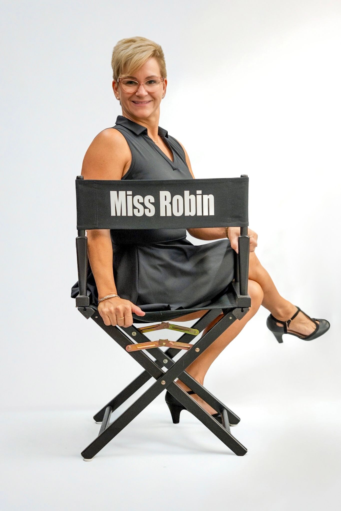 Woman seated on a director’s chair labeled “Miss Robin,” smiling against a white background